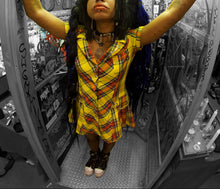 Load image into Gallery viewer, model with dark hair stands inside a graffiti-covered phone booth, wearing a yellow plaid shirt and dark boots, with her arms raised high